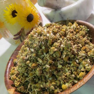 May include: A wooden bowl filled with dried chamomile flowers. The flowers are a light brown color and have a slightly green tint. The bowl is sitting on a white surface.
