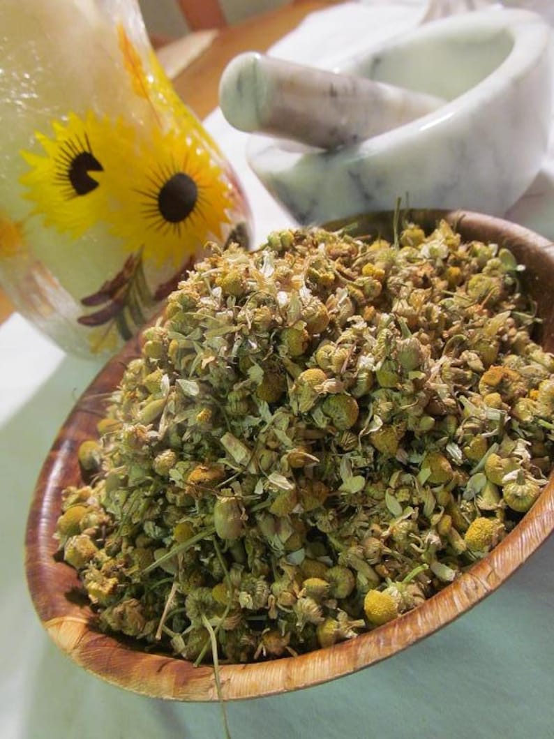 May include: A wooden bowl filled with dried chamomile flowers. The flowers are a light brown color and have a slightly green tint. The bowl is sitting on a white surface.
