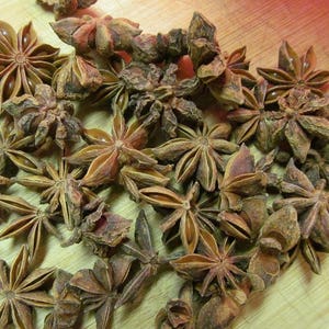 May include: A close-up of a pile of dried star anise pods. The pods are brown and have a star shape with eight points. They are arranged in a random pattern on a wooden surface.