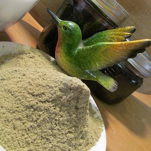 Puede incluir: Un taz&oacute;n blanco lleno de un mont&oacute;n grande de polvo verde claro. Una figurita de colibr&iacute; verde y amarilla est&aacute; posada en el borde del taz&oacute;n.