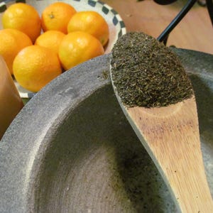 May include: A close-up of a wooden spoon filled with dried green herbs. The spoon is resting on the edge of a gray stone mortar. Several oranges are visible in the background.