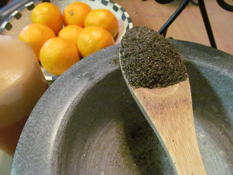 May include: A close-up of a wooden spoon filled with dried green herbs. The spoon is resting on the edge of a gray stone mortar. Several oranges are visible in the background.