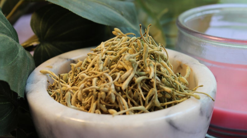 May include: A white marble mortar and pestle with dried, light brown flower buds inside. The mortar and pestle are sitting on a table with green leaves in the background.