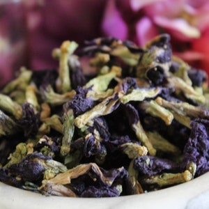 May include: Dried blue butterfly pea flowers in a white bowl. The flowers are a deep blue color with green stems and are ready to be used for tea or other culinary purposes.