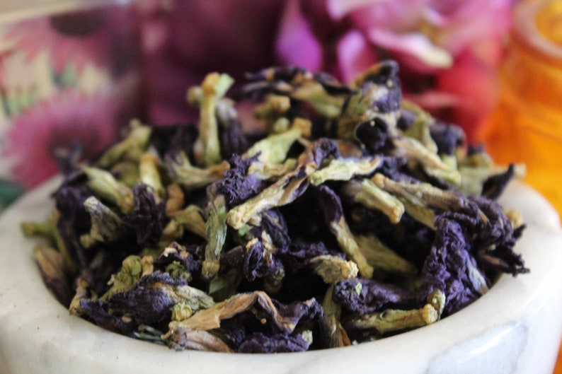 May include: Dried blue butterfly pea flowers in a white bowl. The flowers are a deep blue color with green stems and are ready to be used for tea or other culinary purposes.