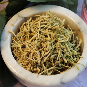 May include: A white marble bowl filled with dried, light brown flower buds. The bowl is sitting on a white surface.