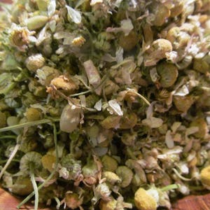 May include: Dried chamomile flowers and leaves in a bowl. The flowers are a pale yellow color and the leaves are a light green.