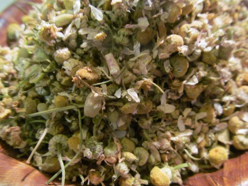 May include: Dried chamomile flowers and leaves in a bowl. The flowers are a pale yellow color and the leaves are a light green.
