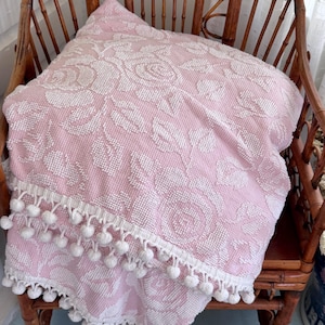 May include: A pink and white floral patterned throw blanket with a white pom-pom trim. The blanket is draped over a wooden chair, showcasing the intricate rose design and textured fabric. The blanket is approximately 50 inches wide.