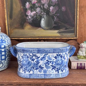 May include: Blue and white ceramic planter with floral and butterfly patterns. The rectangular planter has two curved handles and a detailed design. The color scheme is a classic blue and white, with a traditional aesthetic.