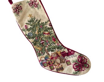 May include: A cream-colored needlepoint Christmas stocking featuring a central green pine tree decorated with ornaments, a star topper, and wrapped gifts at the base. The stocking has a deep red velvet trim and a small hanging loop.