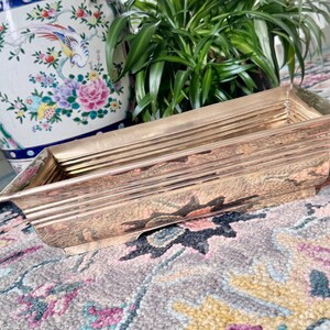 May include: A rectangular, gold-toned metal tray with a textured surface. The tray has a slightly curved shape and is positioned on a patterned rug with floral and geometric designs. A decorative ceramic vase and green plants are in the background.
