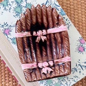 May include: A brown ceramic dish with a ribbed texture, resembling a bundle of sticks, is adorned with pink ribbons and bows. The dish has a rectangular shape and is likely used for decorative purposes.