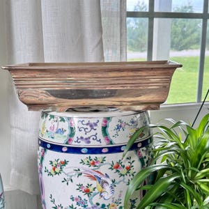 May include: A rectangular, silver-toned metal planter with a ribbed design sits atop a decorative ceramic garden stool. The stool features a colorful floral and bird pattern. The background includes a window and sheer curtains.