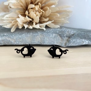 May include: A pair of black pig-shaped stud earrings. Each earring has a heart-shaped cutout and a curled tail. The earrings are displayed on a grey stone surface, with a dried flower and a white background.