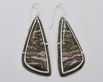 May include: A pair of silver dangle earrings with a teardrop-shaped stone set in a silver frame. The stone is a mix of brown, white, and pink colors with a sparkly texture.