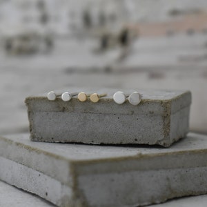 May include: A set of six minimalist stud earrings in silver and gold. The earrings are arranged in a row on a concrete surface.