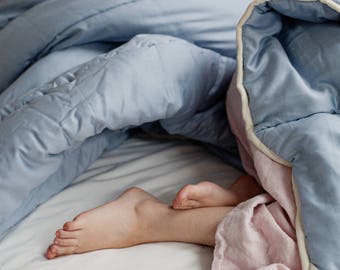 May include: A person's bare feet are tucked under a light blue and pink duvet with a white trim. The duvet is on a white bed.