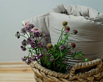 May include: A wicker basket filled with a bouquet of pink and purple wildflowers. A beige comforter is resting on top of the basket. Two gray wool slippers are on the wooden floor.