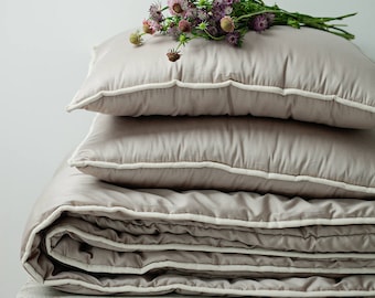 May include: A folded beige duvet with a white trim, and two matching pillows, with a bouquet of purple and pink wildflowers on top.