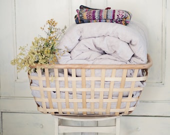 May include: A woven wicker basket filled with a folded white blanket and a patterned throw blanket. The basket is sitting on a white stool in front of a white cabinet.