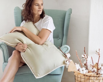May include: A woman sits in a teal armchair holding a light green pillow with white trim. The pillow is made of soft fabric and has a subtle sheen. The woman is wearing a white t-shirt and has long brown hair. There is a basket of cotton branches in the background.