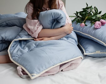 May include: A person sitting on a bed with a light blue duvet and two matching pillows. The person is holding a light blue duvet in their arms. There are pink peonies on the bed.