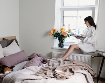 May include: A bedroom with a bed made with a light brown blanket and a few pillows. A person is sitting on a windowsill reading a magazine. There are flowers in a vase on the windowsill.