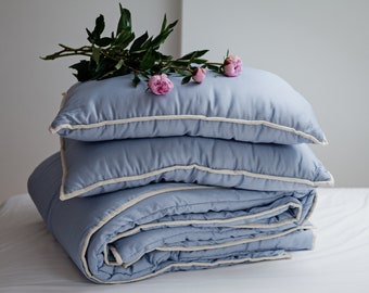May include: A blue quilted comforter with a white trim is folded and stacked with two matching pillows. A sprig of pink peonies rests on top of the pillows.
