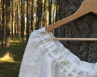 May include: White linen top with a gathered neckline and short sleeves hanging on a wooden hanger in a forest setting.