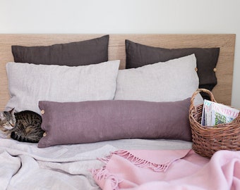 May include: A bed with a brown headboard, a gray linen duvet cover, and a brown linen bolster pillow. There are two gray linen pillows and a pink throw blanket on the bed. A wicker basket with magazines is on the bed next to the bolster pillow. A cat is curled up on the bed.