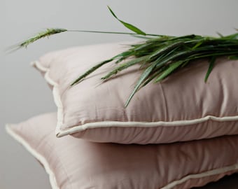 May include: Two pink pillowcases with white piping, stacked on a white surface. A sprig of green grass is resting on top of the pillowcases.