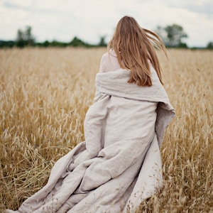 Op de afbeelding: Een vrouw loopt door een veld met hoog, droog gras, draagt een lichtroze shirt en een lange, vloeiende linnen deken.