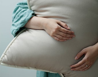 May include: A person holding a large, light beige pillow with a white border. The pillow is made of a soft, smooth fabric.