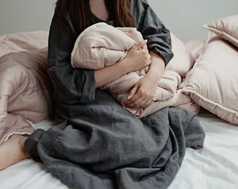 May include: A person in a gray linen dress is sitting on a bed holding a pink pillow. The bed is covered in a pink and white quilt and there are two pink pillows on the bed.