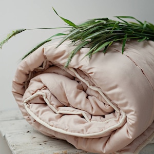 May include: A pale pink duvet with a white trim is folded on a white wooden table. Green grass with seed heads is draped over the duvet.