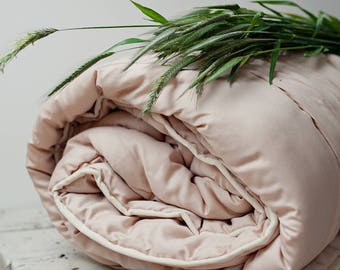 May include: A pale pink duvet with a white trim is folded on a white wooden table. Green grass with seed heads is draped over the duvet.