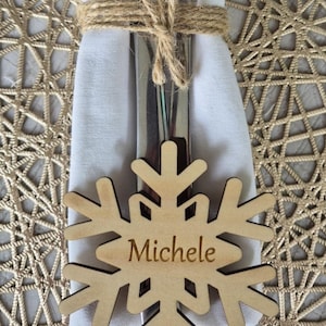 May include: A wooden snowflake place card with the name "Michele" engraved on it. The place card is tied to a white napkin with a fork and knife.
