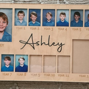 May include: A wooden photo display with a nameplate that reads "Ashley". The display has 12 frames for photos, labelled with the school years "Kindy" through "Year 12".