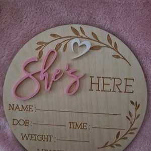May include: A round wooden sign with the words "She's Here" in pink lettering. The sign has a white heart cutout and space to write the baby's name, date of birth, time of birth, weight, and length.