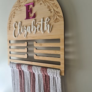 May include: Wooden wall organiser with a pink letter E and the name Elizabeth engraved on it. The organiser has 7 slots for hanging items and a macrame tassel hanging from the bottom.