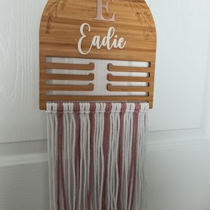 May include: A wooden hanger with a pink and white macrame fringe. The hanger has a cutout arch shape with a rose gold initial "E" and the name "Eadie" written in white script.