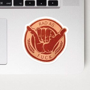 May include: A round sticker with a white border featuring a hand gesture and the words "RAD AS FUCK" in a stylized font. The design is in shades of orange and brown, with a wheat-like border.