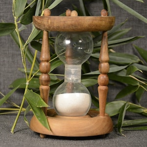 May include: A wooden hourglass with a clear glass bulb filled with white sand. The hourglass is sitting on a gray surface with green bamboo leaves in the background.