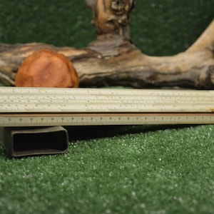 May include: A vintage slide rule, a beige and brown calculating instrument, rests on a dark rectangular support. The slide rule is positioned against a backdrop of green grass, a wooden log, and a brown, rounded object.