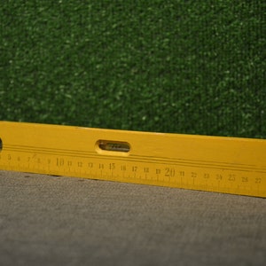 May include: A yellow wooden level with a black and white ruler marked in inches from 1 to 29. The level has a small hole in the center and a clear glass bubble level.