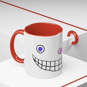 May include: A white ceramic mug with a red rim and handle. The mug features a black and white drawing of a smiling face with two purple eyes.