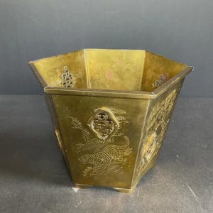 Puede incluir: Una maceta hexagonal de latón con motivos de tigres grabados y paneles decorativos calados. La superficie metálica muestra una pátina desgastada con grabados visibles de tigres entre pinos en cada uno de los seis lados del recipiente.