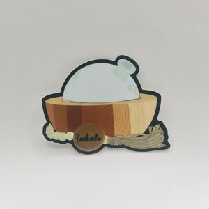 May include: A sticker illustration of a white dome-shaped diffuser sitting in a wooden bowl with varying brown tones. A string of light-colored beads with a brown circular tag featuring the word Inhale in black script and a tassel are attached.