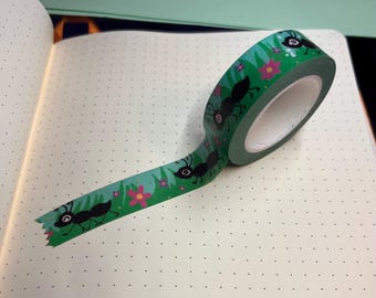 Op de afbeelding: Een rol decoratieve washi-tape met een herhalend patroon van zwarte mieren, roze bloemen en groen gras op een turquoise achtergrond. De tape is uitgerold op een notitieboek met stippen.